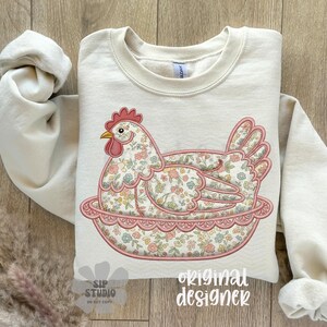 May include: Cream-colored sweatshirt featuring an embroidered hen design. The hen is resting in a pink floral nest, with a red comb and yellow beak. The hen and nest are decorated with a floral pattern. The words "original designer" are printed on the bottom.