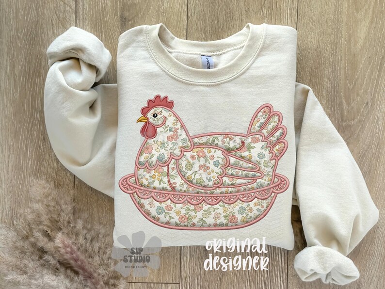 May include: Cream-colored sweatshirt featuring an embroidered hen design. The hen is resting in a pink floral nest, with a red comb and yellow beak. The hen and nest are decorated with a floral pattern. The words "original designer" are printed on the bottom.
