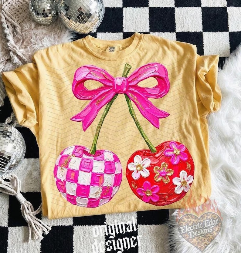 May include: A yellow t-shirt featuring a large graphic of two cherries tied with a bright pink bow. One cherry has a pink and white checkered pattern, while the other is red with white and pink flowers. The design has a textured, painted appearance.