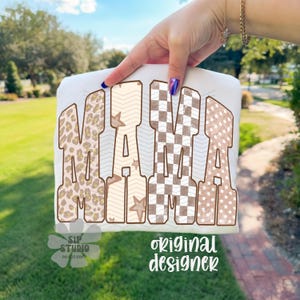 May include: White t-shirt with the word "MAMA" in large, decorative letters. The letters feature various patterns, including leopard print, chevron, checkered, and star designs. The words "SIP STUDIO DO NOT COPY" and "original designer" are also visible.
