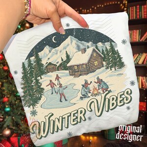 May include: A white t-shirt featuring a graphic print of a snowy mountain landscape with log cabins, pine trees, a crescent moon, and people ice skating and sitting by a campfire. The text "WINTER VIBES" is printed in a large, retro-style font.