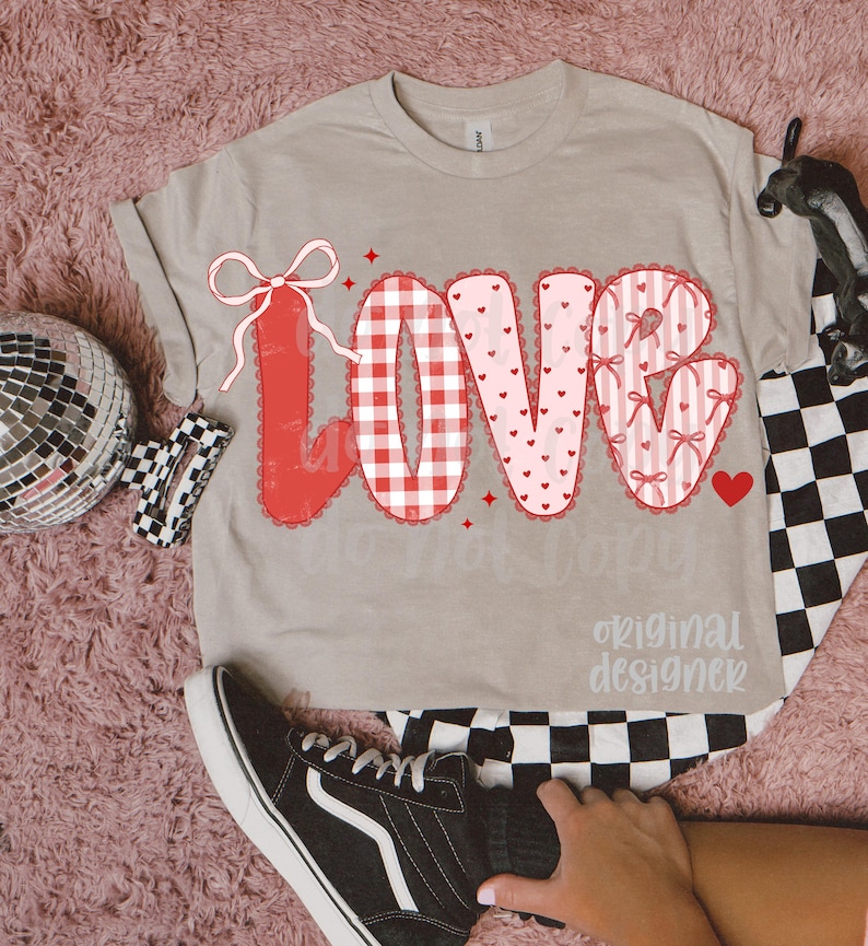 May include: A beige t-shirt with the word "LOVE" in a red and pink design. The "L" is a red bow, the "O" is a red and white gingham pattern, the "V" is pink with hearts, and the "E" is pink with bows. The shirt has the text "original designer".
