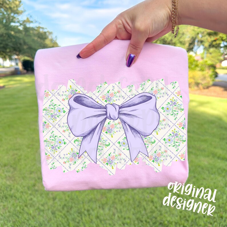 May include: A light pink t-shirt featuring a large lavender bow design. The bow is centered on a patterned patch with floral and diamond elements. The words "original designer" are written in white script on the bottom right.