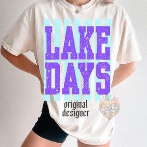 K&ouml;nnte beinhalten: Cremefarbenes T-Shirt mit den Worten "LAKE DAYS" in gro&szlig;en, verwaschenen lila Buchstaben. Das Design umfasst hellblaue vertikale Streifen und den Text "original designer" in einer gotischen Schriftart. Ein kleines Logo ist rechts sichtbar.