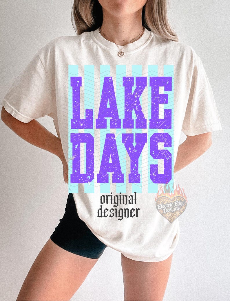 K&ouml;nnte beinhalten: Cremefarbenes T-Shirt mit den Worten "LAKE DAYS" in gro&szlig;en, verwaschenen lila Buchstaben. Das Design umfasst hellblaue vertikale Streifen und den Text "original designer" in einer gotischen Schriftart. Ein kleines Logo ist rechts sichtbar.
