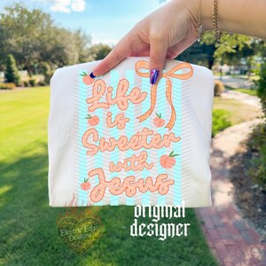 May include: White t-shirt with the text "Life is Sweeter with Jesus" in peach-colored script, accented with peach illustrations and a light blue chevron pattern. The design includes a peach-colored bow and the words "original designer".