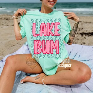 May include: A mint green t-shirt with the words "LAKE BUM" in pink, set against a teal leopard print background. The shirt is a casual style, perfect for a day at the lake or beach.