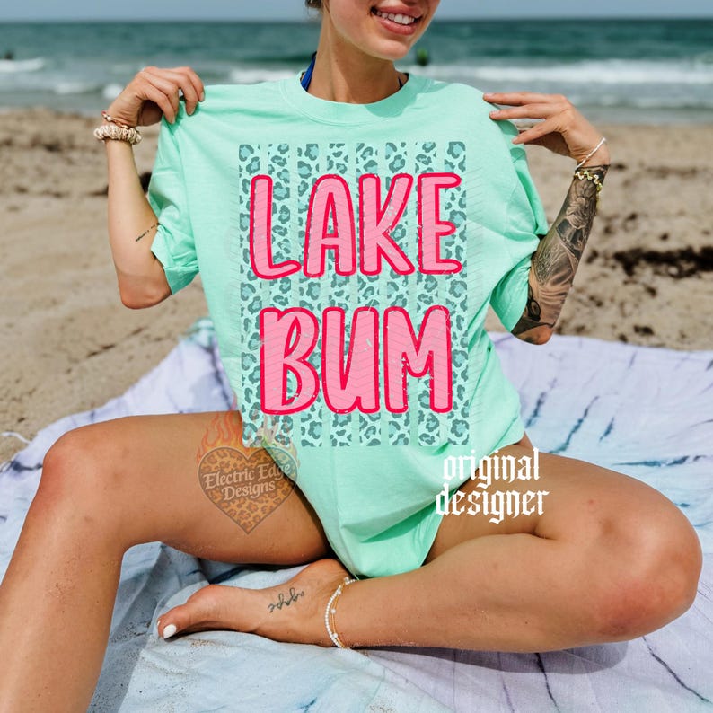May include: A mint green t-shirt with the words "LAKE BUM" in pink, set against a teal leopard print background. The shirt is a casual style, perfect for a day at the lake or beach.