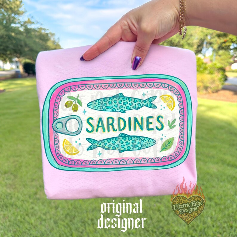May include: A light pink t-shirt featuring a sardine can design. The design includes two turquoise fish, olives, lemon slices, and the word "SARDINES" in white. The shirt has the text "original designer" at the bottom.