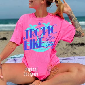 May include: A bright neon pink short-sleeve t-shirt featuring a large graphic print in blue and light blue. The design includes two palm trees and the text "TROPIC LIKE It's Hot" in a bold, stylized font. Small flower icons accent the text.