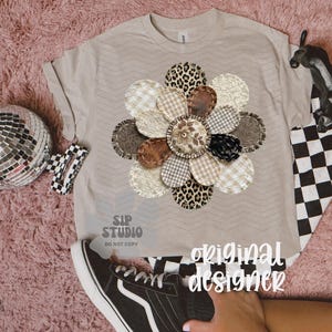 May include: A beige t-shirt with a large, appliqued flower design. The flower features various patterned petals, including leopard print, gingham, and textured fabrics. The words "original designer" are visible on the shirt.