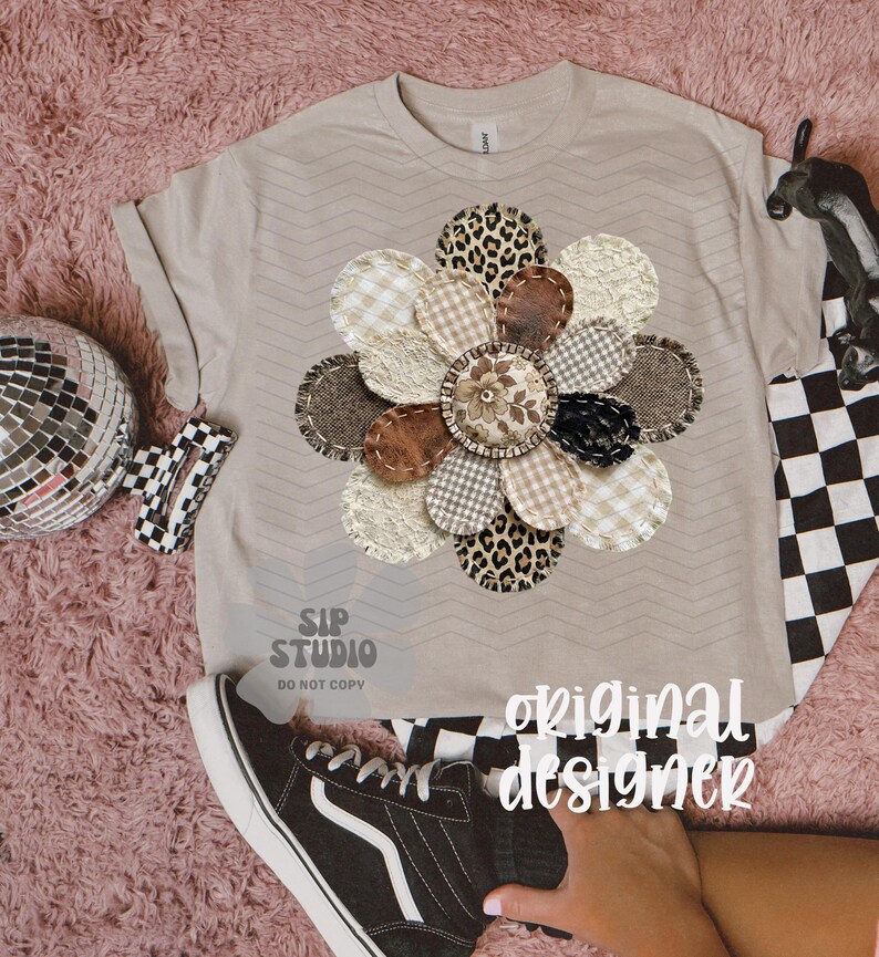 May include: A beige t-shirt with a large, appliqued flower design. The flower features various patterned petals, including leopard print, gingham, and textured fabrics. The words "original designer" are visible on the shirt.