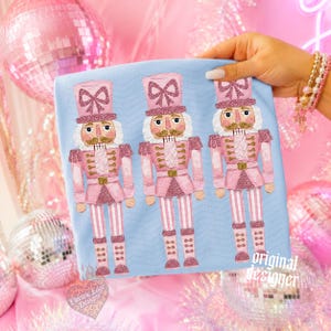 May include: A light blue t-shirt featuring a repeating graphic print of three pink nutcracker figures. Each nutcracker wears a tall pink hat with a glittery bow, a pink uniform with gold accents, and pink striped trousers. The fabric has a subtle chevron pattern.