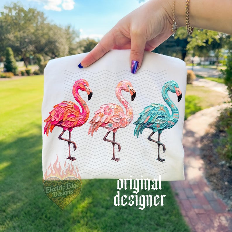 May include: White sweatshirt featuring three colorful flamingo designs. The flamingos are in shades of pink, coral, and teal, with intricate patterns. The text "Electric Edge Designs" and "original designer" are also visible.