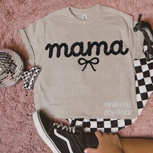 May include: A beige t-shirt with the word "mama" in black, cursive, rope-like letters, and a black bow. The text "original designer" is printed in a light gray color. A black and white checkered pattern is visible.