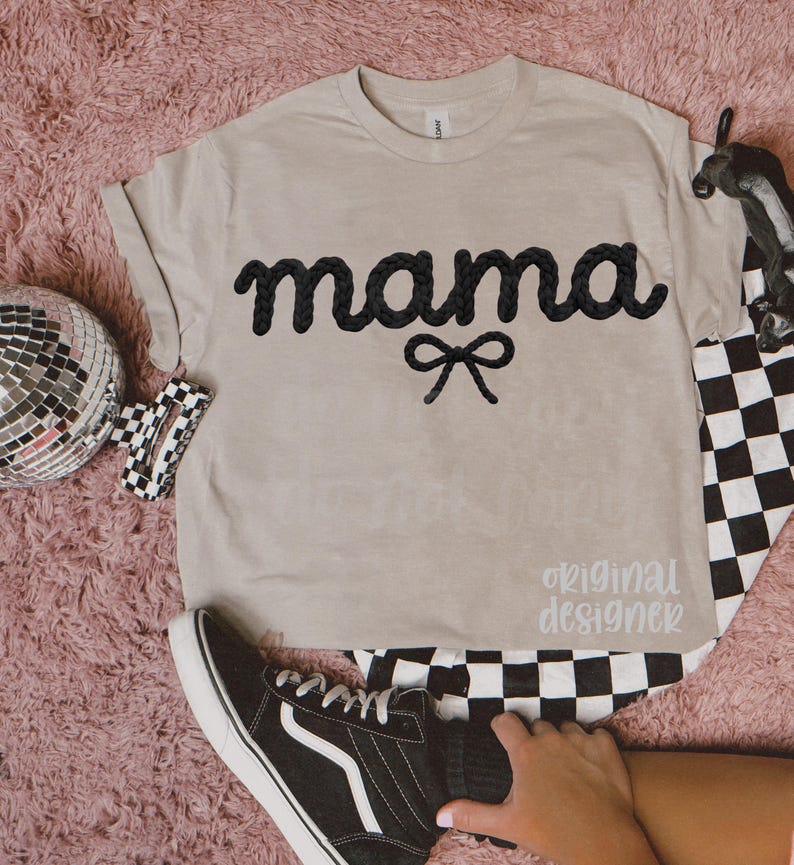 May include: A beige t-shirt with the word "mama" in black, cursive, rope-like letters, and a black bow. The text "original designer" is printed in a light gray color. A black and white checkered pattern is visible.
