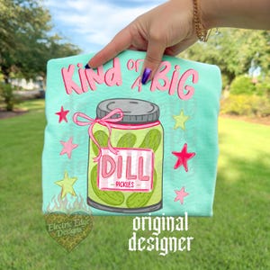 May include: A light teal t-shirt with the text "Kind of a BIG" in pink script. A jar of dill pickles is in the center, with pink stars and a pink bow. The words "DILL - PICKLES" are on the jar. The words "original designer" are at the bottom.