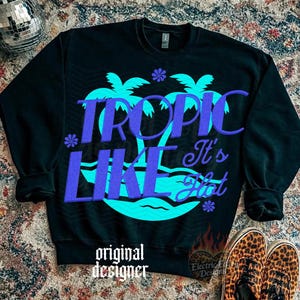 May include: A black long-sleeve sweatshirt featuring a large graphic of two turquoise palm trees and wavy lines. The text "TROPIC LIKE It's Hot" is printed in a bold purple font across the center. Small purple flower icons are scattered around.