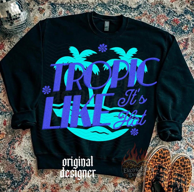 May include: A black long-sleeve sweatshirt featuring a large graphic of two turquoise palm trees and wavy lines. The text "TROPIC LIKE It's Hot" is printed in a bold purple font across the center. Small purple flower icons are scattered around.