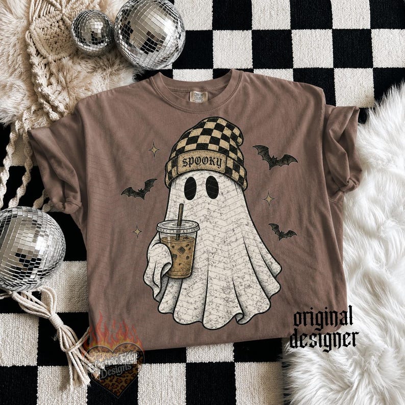 May include: A brown t-shirt featuring a graphic of a white ghost wearing a tan and black checkered beanie with the word SPOOKY printed on it. The ghost holds an iced coffee, surrounded by small black bats and star shapes on the fabric.