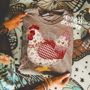May include: A tan t-shirt with a patchwork chicken design. The chicken has a red comb, beak, and wattle, with a body of white floral and red gingham patterns. The text "original designer" is visible on the shirt.