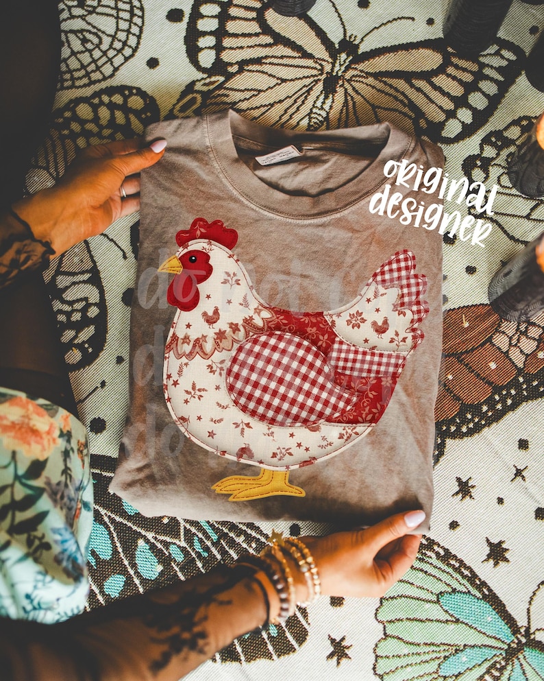 May include: A tan t-shirt with a patchwork chicken design. The chicken has a red comb, beak, and wattle, with a body of white floral and red gingham patterns. The text "original designer" is visible on the shirt.