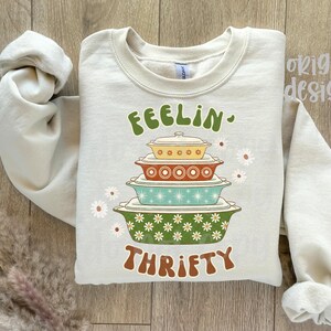 Pu&ograve; includere: Una felpa beige chiaro con le parole "FEELIN' THRIFTY" in lettere marroni e verdi. La grafica presenta una pila di pirofile colorate. Accenti di margherite bianche sono sparsi intorno al design.