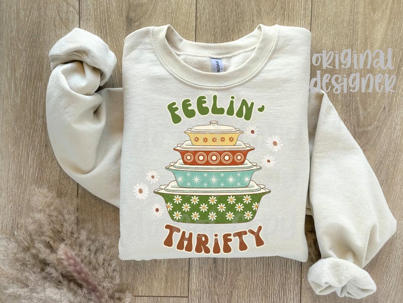 Pu&ograve; includere: Una felpa beige chiaro con le parole "FEELIN' THRIFTY" in lettere marroni e verdi. La grafica presenta una pila di pirofile colorate. Accenti di margherite bianche sono sparsi intorno al design.