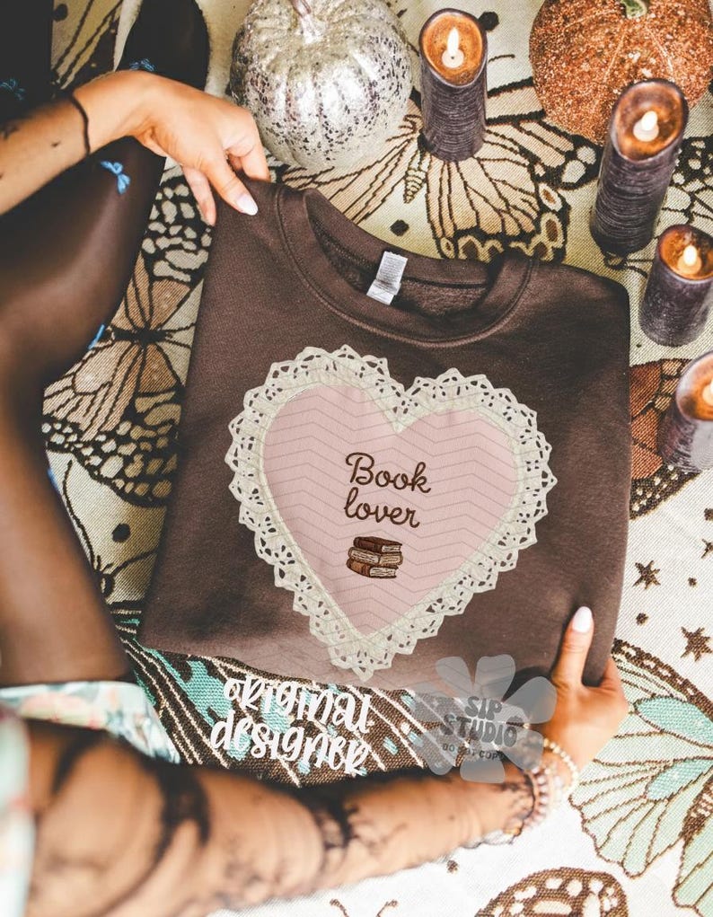 May include: A brown sweatshirt featuring a pink heart design with the words "Book lover" and a stack of books. The heart is trimmed with white lace. The sweatshirt is held by a person.