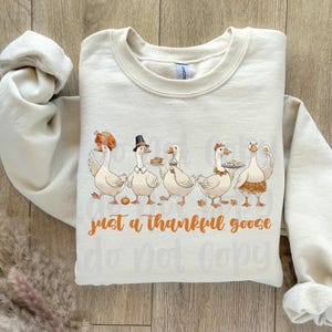 May include: Cream-colored sweatshirt with a Thanksgiving-themed graphic. The design features five cartoon geese, each holding food items like a turkey leg and pie. The text "just a thankful goose" is written in orange script.
