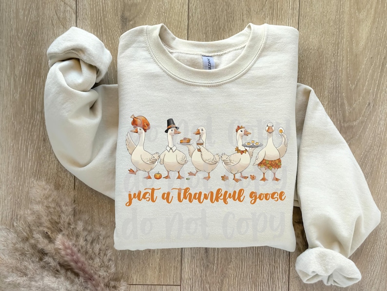 May include: Cream-colored sweatshirt with a Thanksgiving-themed graphic. The design features five cartoon geese, each holding food items like a turkey leg and pie. The text "just a thankful goose" is written in orange script.