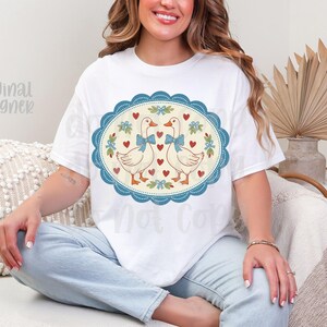 May include: White t-shirt featuring a whimsical design of two geese with blue bow ties, surrounded by hearts and floral accents within a blue oval frame. The words "Original Designer" are visible on the shirt.