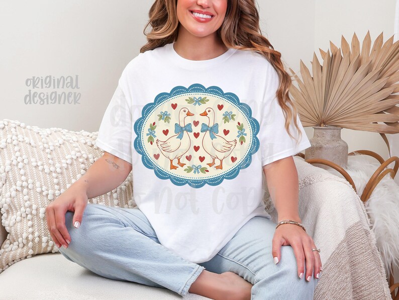 May include: White t-shirt featuring a whimsical design of two geese with blue bow ties, surrounded by hearts and floral accents within a blue oval frame. The words "Original Designer" are visible on the shirt.