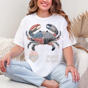 May include: White t-shirt featuring a patchwork crab design in red, blue, and white patterns. The shirt includes the text "original designer" and a heart-shaped logo with text.