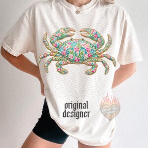 K&ouml;nnte beinhalten: Cremefarbenes T-Shirt mit einem detaillierten Krabben-Design mit floralen Mustern in Rosa, Blau und Gr&uuml;n. Das Shirt hat den Text "original designer" in schwarzer gotischer Schrift. Ein herzf&ouml;rmiges Logo mit dem Text "Electric Edge Designs" ist ebenfalls sichtbar.