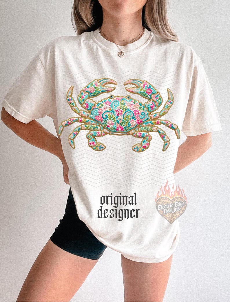 K&ouml;nnte beinhalten: Cremefarbenes T-Shirt mit einem detaillierten Krabben-Design mit floralen Mustern in Rosa, Blau und Gr&uuml;n. Das Shirt hat den Text "original designer" in schwarzer gotischer Schrift. Ein herzf&ouml;rmiges Logo mit dem Text "Electric Edge Designs" ist ebenfalls sichtbar.