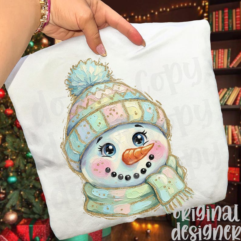 May include: A white t-shirt featuring a colorful, textured illustration of a snowman wearing a pastel-colored knit hat with a pom-pom and a matching scarf. The snowman has large blue eyes, a carrot nose, and a dotted smile, with gold accents.