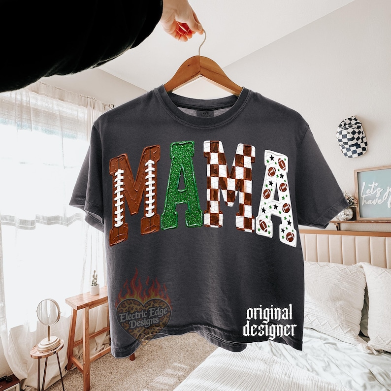 May include: A charcoal gray t-shirt featuring the word MAMA in large letters. The M is brown with football laces, the A is green glitter, the M is a brown and white checkerboard pattern, and the A is white with footballs and stars.