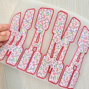 Op de afbeelding: Bloemen warmteoverdracht vinyl met het woord "MAMA" in een vetgedrukte, blokletter lettertype. De letters zijn gevuld met een roze bloemenpatroon en rood omlijnd. Het vinyl wordt door een hand vastgehouden.