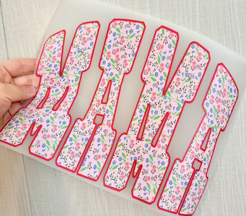 Op de afbeelding: Bloemen warmteoverdracht vinyl met het woord "MAMA" in een vetgedrukte, blokletter lettertype. De letters zijn gevuld met een roze bloemenpatroon en rood omlijnd. Het vinyl wordt door een hand vastgehouden.