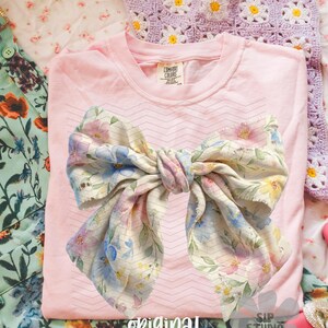 May include: A light pink t-shirt with a large bow design featuring blue, pink, and yellow floral patterns. The words "original designer" are printed at the bottom. The shirt is made by Comfort Colors.