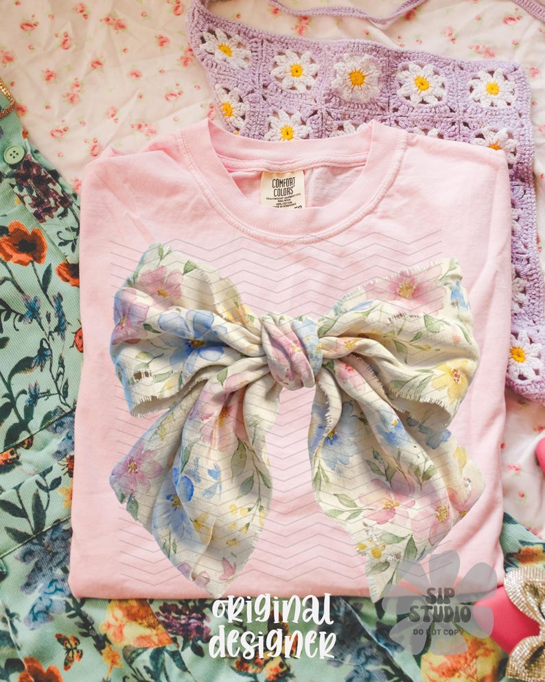 May include: A light pink t-shirt with a large bow design featuring blue, pink, and yellow floral patterns. The words "original designer" are printed at the bottom. The shirt is made by Comfort Colors.