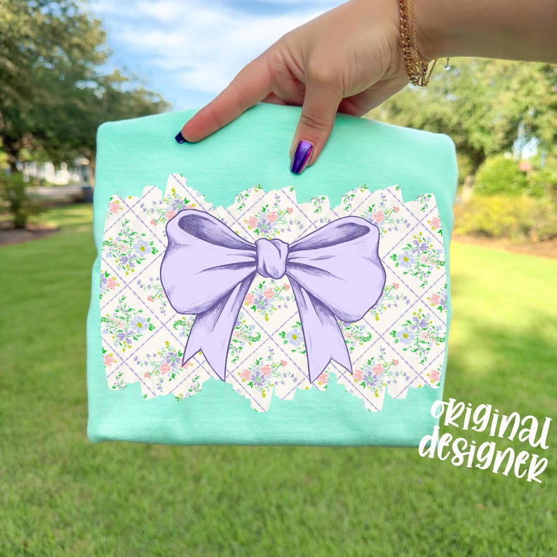 May include: A mint green t-shirt featuring a large lavender bow design on a floral patterned background. The design is framed by a decorative border. The words "original designer" are written in a script font on the lower right corner.