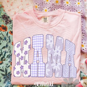 May include: A light pink Comfort Colors t-shirt with the word "MAMA" in large, block letters. The letters feature floral, gingham, and striped patterns in shades of purple and white. The shirt is on a floral patterned surface.