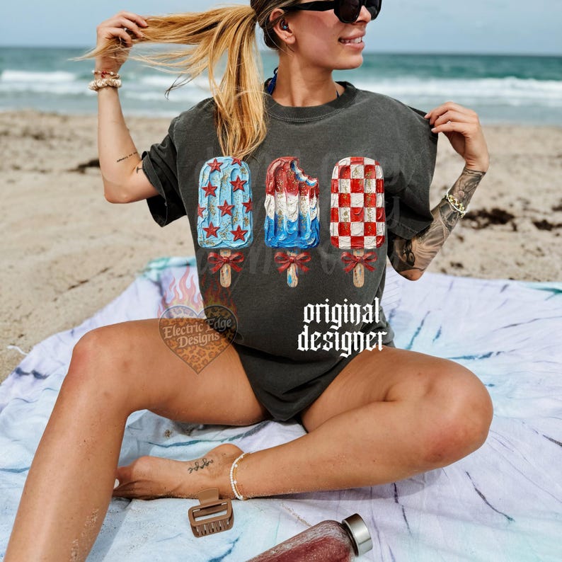 Pu&ograve; includere: T-shirt grigio scuro con tre disegni di ghiaccioli rossi, bianchi e blu, ognuno con un fiocco. Il testo "original designer" &egrave; stampato sulla maglietta. L'immagine &egrave; scattata su una spiaggia.
