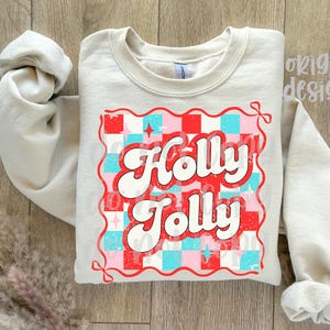 May include: A light beige sweatshirt with the words "Holly Jolly" in a retro font. The text is set against a red, pink, and blue checkered pattern, with a red border. The sleeves are rolled up.