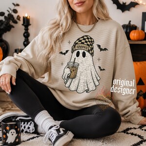 May include: A beige crewneck sweatshirt featuring a graphic of a ghost wearing a black and tan checkered beanie, holding an iced coffee, with the text SPOOKY on the beanie. The ghost is surrounded by small black bats and star-shaped accents.