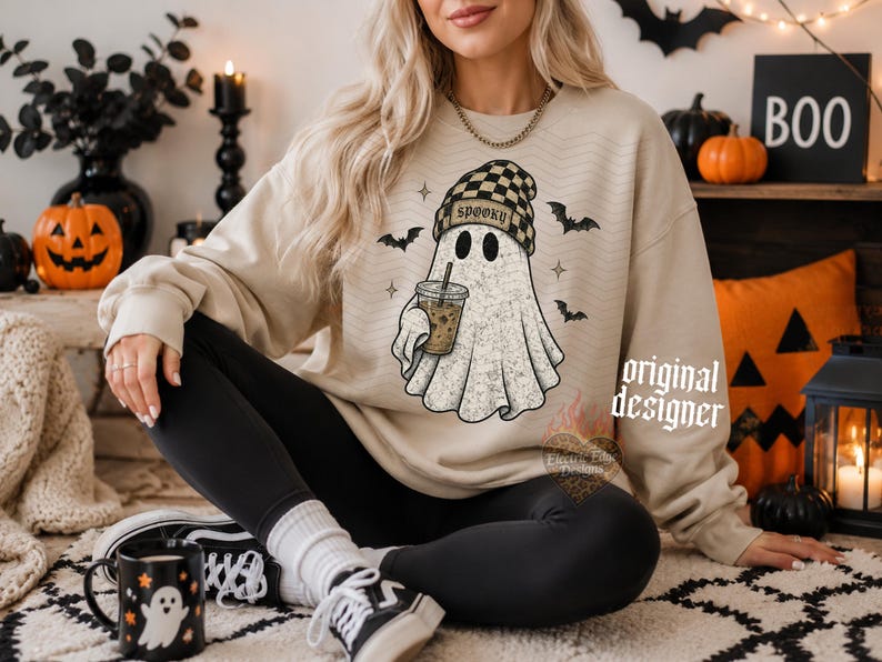 May include: A beige crewneck sweatshirt featuring a graphic of a ghost wearing a black and tan checkered beanie, holding an iced coffee, with the text SPOOKY on the beanie. The ghost is surrounded by small black bats and star-shaped accents.