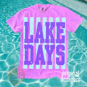 May include: A lilac-colored t-shirt with the words "LAKE DAYS" in purple block letters. The text is set against a backdrop of light blue vertical stripes. The shirt has a crew neck and short sleeves. The words "original designer" are printed on the bottom right.