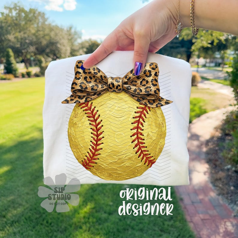Op de afbeelding: Wit T-shirt met een softbal ontwerp. De softbal is geel met rode stiksels en een luipaardprint strik. De tekst "original designer" is zichtbaar.